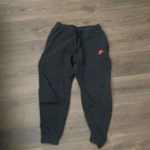 Nike Tech Fleece Byxor  - Nypris 1100kr. De är knappt använda. Svarta mjukisbyxor från Nike med orange logga på vänster lår. Byxorna har resår i midjan och vid bensluten samt dragkedjeförsedda fickor på sidorna. Perfekta för chill eller träning.
