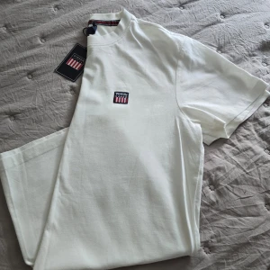 Vit t-shirt från Vinson - Säljer en fräsch vit t-shirt från Vinson i modellen Massimo. T-shirten har ett broderat Vinson-märke på bröstet och klassisk rund hals med randig detalj på insidan. Perfekt basic-plagg i mjuk bomull.