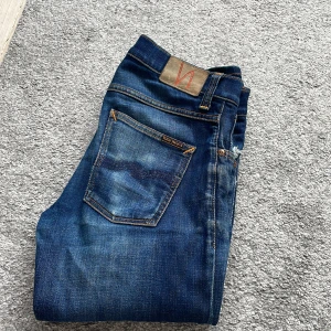 Blå jeans från Nudie Jeans Co - Säljer ett par klassiska blå jeans från Nudie Jeans Co med slitningar och orangea sömmar. Jeansen har fem fickor och normal passform. Perfekta för dig som gillar en avslappnad stil.