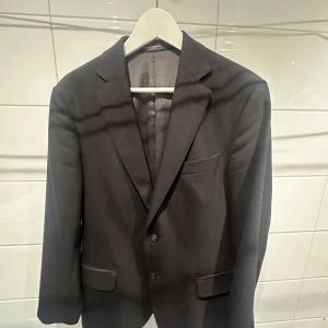 Svart kavaj från Oscar Jacobson - Klassisk svart kavaj från Oscar Jacobson med två knappar framtill och fickor med lock!🙌 Kavajen har en stilren design och är helfodrad med slät insida. Perfekt nu inför sommaren! Passar perfekt till en vit tisha eller skjorta!!🫱🏻‍🫲🏼