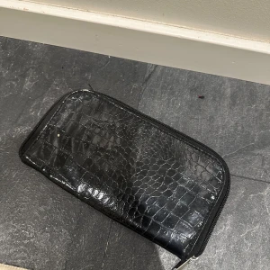Svart borstplånbok med mönster - Säljer en svart borstplånbok med krokodilpräglat mönster och dragkedja runtom. Plånboken har flera fack på insidan för borstar! Den har lite smink spår på insidan men går säkert att tvätta bort! 