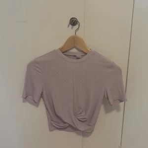 Ljuslila croppad ribbad topp - Säljer en ljuslila croppad topp med korta ärmar och rund halsringning. Toppen är ribbad och har en snygg twist-detalj framtill som ger en cool look. Perfekt att matcha med höga jeans eller kjol.
