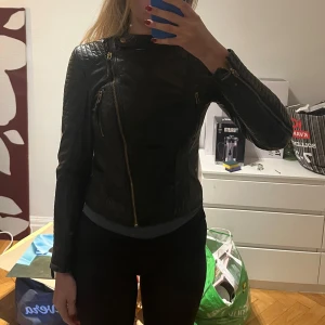 Svart skinnjacka med asymmetrisk dragkedja - Säljer en svart skinnjacka med cool asymmetrisk dragkedja och detaljerade sömmar på axlarna. Jackan har ståkrage med tryckknapp och guldiga metalldetaljer. Perfekt för en edgy look. Äkta läder från märket primeboots 