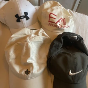 Fyra kepsar från olika märken - Skriv om du är intresserad av någon så snackar vi pris Fyra kepsar i olika färger och märken: vit Under Armour med svart logga, vit Emporio Armani med röd logga, beige keps med liten broderad detalj framtill och en grå nike med vit swoosh  