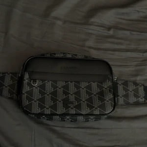 Lacoste Waist Bag - Tar emot byten framför allt! Säljer en svart axelväska från Lacoste med geometriskt vitt mönster och justerbart band med metallspänne. Väskan har dragkedja. Insidan är fodrad i grått och orange. Perfekt för att bära det viktigaste. 