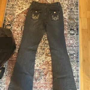 Grå bootcut jeans med broderade bakfickor - Säljer ett par gråa bootcut jeans med snygga broderade detaljer på bakfickorna. Jeansen har klassisk femficksdesign och stängs med knapp och dragkedja. Perfekta för dig som gillar en retroinspirerad look.