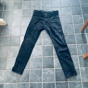 Svarta jeansbyxor från Replay Anbass - Säljer ett par svarta Replay Anbass jeans med klassisk femficksdesign och raka ben. Jeansen har normal passform och är tillverkade i ett mjukt denimtyg som sitter snyggt. Perfekta för dig som gillar stilrena svarta jeans.