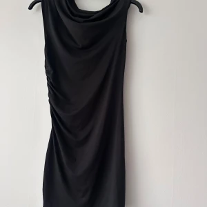 Svart draperad klänning från Divided - Säljer en svart klänning från Divided med snygg drapering och cowlneck. Klänningen är ärmlös och har en tight passform som framhäver siluetten. Perfekt för dig som gillar minimalistisk stil.