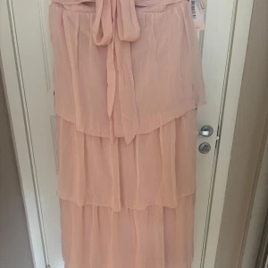 Ljusrosa långklänning med volanger - Superfin ljusrosa långklänning med flera lager volanger och knytband i midjan. Perfekt för en bal,fest eller för sommaren. Klänningen är luftig och bekväm. Klänningen är S men kan även passa som XS