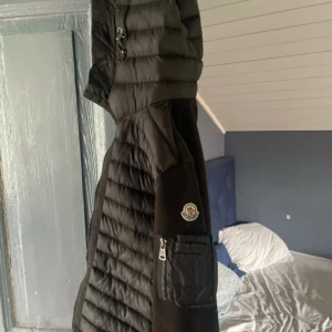 Svart pufferjacka med huva från Moncler - Säljer en svart pufferjacka från Moncler med huva och dragkedja. Jackan har en quiltad design, ficka med dragkedja på ärmen och klassisk Moncler-logga. Perfekt för alla stilar. Den har inga skador eller sånt skulle säga att den är i ny skick
