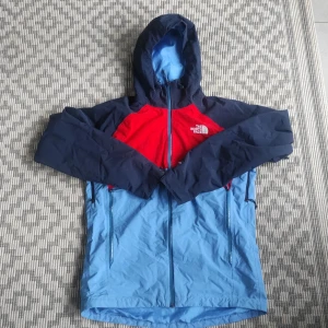 Blå och röd vindjacka från The North Face - Säljer en snygg vindjacka från The North Face i blått och rött. Jackan har huva, dragkedja framtill och två sidofickor med dragkedja. Perfekt för utomhusaktiviteter och är tillverkad i 100% polyester.