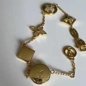 Guldfärgat armband från Luis Vitton med kristalldetaljer - Exklusivt armband i guldfärg från LV med flera ikoniska berlocker, vissa dekorerade med gnistrande kristaller. Armbandet har LV-logga och olika former som fyrklöver och blomma. Perfekt för dig som vill ha något lyxigt och unikt.