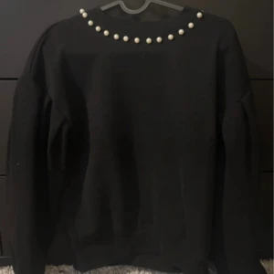 Svart sweatshirt med pärlor från Kappahl - Svart sweatshirt från Kappahl i storlek S. Tröjan har rund halsringning och är dekorerad med pärlor längs halsen för en snygg detalj. Perfekt för dig som vill ha något enkelt men ändå med en twist.