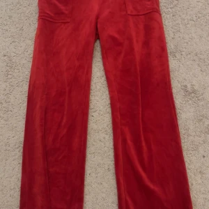 Röda velourbyxor från Juicy Couture - Säljer ett par klassiska röda velourbyxor från Juicy Couture med raka ben och fickor både fram och bak. Byxorna har en mjuk och glansig finish som ger en lyxig känsla. Perfekta för en avslappnad och trendig look.
