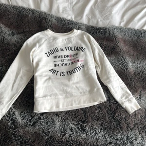 Vit sweatshirt från Zadig & Voltaire - Säljer en vit sweatshirt från Zadig & Voltaire med svart tryck framtill och en liten rosa detalj. Tröjan har rund halsringning och långa ärmar. Perfekt för en cool och avslappnad stil.