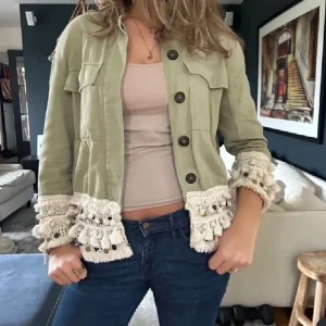 Grön jacka med virkade detaljer - Snygg grön jacka med stora virkade och volangprydda detaljer i beige vid ärmslut och nederkant. Jackan har stora knappar framtill och två bröstfickor. Perfekt för dig som vill sticka ut med en unik stil.