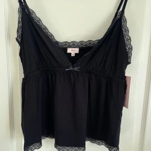 Svart spetslinne - Säljer ett svart linne från Nelly med spetsdetaljer längs kanterna och en liten rosett framtill. Linnet har smala axelband och v-ringning. Aldrig använd🩷
