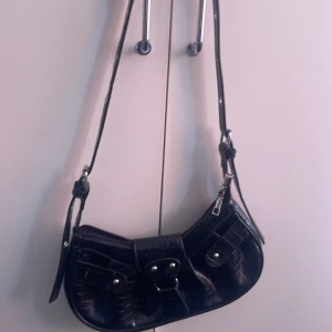 Svart axelväska med krokodilpräglat mönster - Säljer en svart axelväska med krokodilpräglat mönster och silverfärgade detaljer. Väskan har justerbar axelrem, dragkedja upptill och dekorativa spännen framtill. Perfekt för dig som vill ha en trendig accessoar till din outfit.