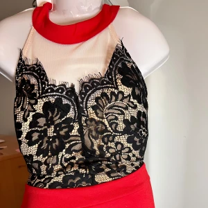 Röd klänning med spetsdetaljer ärmlös  - Säljer en snygg röd klänning med svart blommig spets upptill och hög hals. Klänningen har en vid kjol och är ärmlös, perfekt för festliga tillfällen. Stängs med dragkedja i ryggen och har en elegant design.