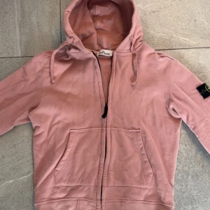 Rosa hoodie från Stone Island - Säljer en rosa hoodie från Stone Island med dragkedja och klassisk logga på ärmen. Tröjan har huva med snörning och två stora fickor framtill. Perfekt för en avslappnad och stilren look.