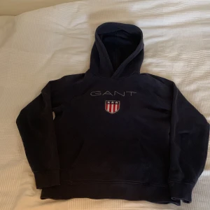 Mörkblå hoodie från GANT - Säljer en mörkblå hoodie från GANT med broderad logga och sköld på bröstet. Tröjan har huva och klassisk passform, perfekt för en avslappnad stil. Mjuk och bekväm med ribbade muddar vid ärmslut och nederkant.