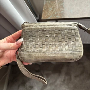 Grå flätad plånbok med handledsrem - Säljer en grå plånbok i skinn med flätad framsida och dragkedja. Plånboken har flera fack för kort och en smidig handledsrem. Perfekt för att hålla ordning på det viktigaste.