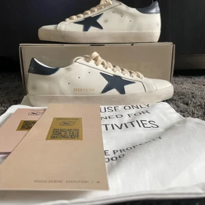 Golden Goose Superstar sneakers med blå stjärna - Golden Goose Superstar sneakers i vitt skinn med mörkblå stjärndetalj på sidan och matchande blå hälkappa. Klassisk rund tå, platt sula och beige snörning. Märkeslogga på hälen och insidan. Perfekta för en trendig streetstyle.