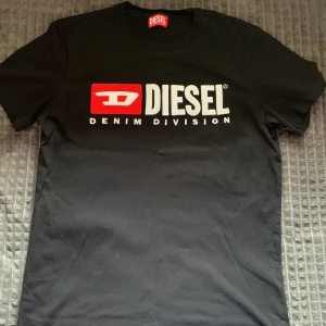 Svart t-shirt från Diesel med tryck - Snygg svart t-shirt från Diesel med stort vitt och rött tryck på bröstet där det står 'Diesel Denim Division'. Klassisk rund hals och korta ärmar. Perfekt för dig som gillar streetwear och vill ha en stilren look. Säljer den då den inte kommer till användning. Den är i princip helt ny