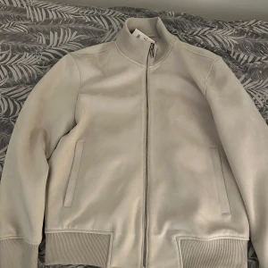 Beige bomberjacka från ZARA - Säljer en stilren beige bomberjacka från ZARA i storlek M. Jackan har dragkedja framtill, ribbade muddar vid ärmslut och midja samt två sidofickor. Perfekt för dig som gillar en clean och modern look. EJ ANVÄND