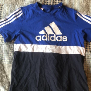 Blå och svart t-shirt från Adidas - Snygg t-shirt från Adidas i blått, vitt och svart med klassiska ränder på axlarna och stor logga framtill. T-shirten har rund hals och korta ärmar. Perfekt för en sportig look.