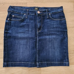 Mörkblå jeanskjol från Lee - Säljer en klassisk mörkblå jeanskjol från Lee med fem fickor och knappstängning framtill. Kjolen har en rak passform och är i kraftigt denimtyg. Perfekt till sneakers eller boots för en avslappnad look.