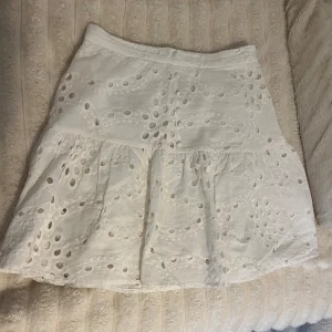 Vit broderad kjol med volang - Superfin vit kjol med broderade detaljer och volang. Kjolen har hålmönster och är perfekt för varma dagar. Den är kort och har en luftig känsla, vilket gör den extra skön att bära. 