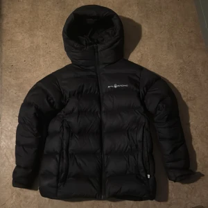 Svart dunjacka från Sail Racing - Säljer en svart dunjacka från Sail Racing med huva och dragkedja. Jackan är vadderad med 600 fill power dun och har två sidofickor med dragkedja. Perfekt för kalla dagar.