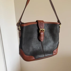Svart och brun axelväska med spänne - Säljer en stilren svart axelväska med bruna detaljer och axelrem. Väskan har ett dekorativt spänne framtill och är tillverkad i skinn med struktur. Köpt second hand och använd fåtal gånger✨