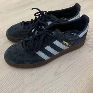 Adidas Spezial sneakers i mörkblå mocka - Säljer ett par Adidas Spezial sneakers i mörkblå mocka med vita ränder och gummisula. Skorna har snörning och guldfärgad Spezial-text på sidan. Klassisk retrostil med rund tå och Adidas-logga på innersulan.