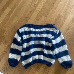 Blåvit randig stickad tröja från Design by Si - Mysig stickad tröja med breda blå och vita ränder från Design S. Tröjan har lång ärm och en avslappnad passform med ribbade muddar vid ärmslut och nederkant. Liten skada vid axeln som bilden visar