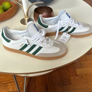Adidas Samba OG vita och gröna sneakers - Säljer ett par klassiska Adidas Samba OG sneakers i vitt skinn med gröna ränder och detaljer. Skorna har gummisula, snörning och blå Adidas-logga på plösen. Perfekta för dig som gillar retrostil och streetwear. Helt oanvända lapp kvar från butik