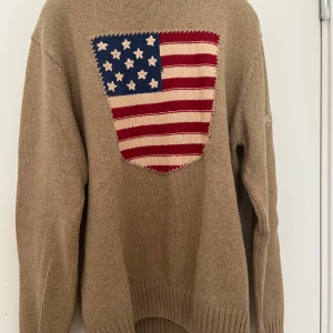 Beige stickad tröja med amerikansk flagga från Gant - Säljer en beige stickad tröja från Gant med en stor amerikansk flagga framtill. Tröjan har långa ärmar och ribbade muddar vid ärmslut och nederkant. Broderad Gant-signatur på ena ärmen. 100% bomull.