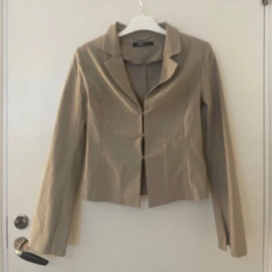 Beige kavaj med klassisk krage - Säljer en beige kavaj från Zara med klassisk krage och lång ärm. Kavajen har en figurnära passform och stängs framtill med dolda knappar. Perfekt för en stilren look och enkel att matcha med olika outfits.