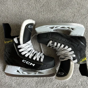 CCM Super Tacks skridskor - Säljer ett par svarta CCM Super Tacks skridskor med vita snören och detaljer i gult. Skridskorna har en robust konstruktion med kolfibermönster på sidan och vit plast under skenan. Perfekta för dig som vill ha grym kontroll på isen.