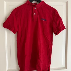 Röd pikétröja från Sebago - Röd pikétröja från Sebago med klassisk krage och två knappar framtill. Tröjan har korta ärmar och är tillverkad i 100% bomull. Liten broderad logga på bröstet. Perfekt för en avslappnad och stilren look.