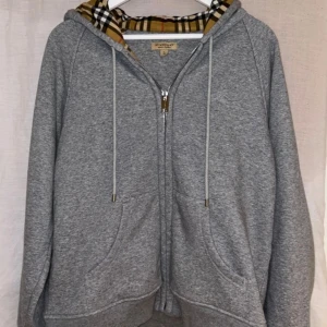 Grå hoodie från Burberry - Säljer en stilren grå hoodie från Burberry med klassiskt rutigt mönster i huvan. Tröjan har dragkedja och broderad logga på bröstet. Perfekt för en avslappnad och trendig look. Liten i storleken är en XL men sitter mer som en L