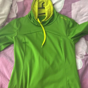 Grön hoodie från Quechua - Säljer en grön hoodie från Quechua med kontrasterande ljusgul huva och snörning. Tröjan har lång ärm och är tillverkad i ett syntetmaterial som passar perfekt för utomhusaktiviteter.