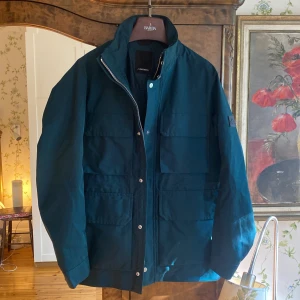 J.Lindeberg fieldjacket - Säljer en mörkblå jacka från J.Lindeberg i storlek M. Jackan har flera stora fickor framtill, dragkedja och tryckknappar. Klassisk och stilren design med lång ärm och hög krage. Perfekt för dig som vill ha en snygg och funktionell jacka. Funkar för alla årstider. Helt ny utan prislapp, aldrig använd. Färgen är mörk grön. 
