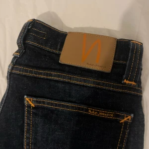 Mörkblå jeans från Nudie Jeans - Säljer ett par mörkblå jeans från Nudie Jeans med klassiska orange sömmar och läderpatch bak. Modellen har fem fickor och raka ben. Perfekta för dig som gillar stilrena och tidlösa jeans.