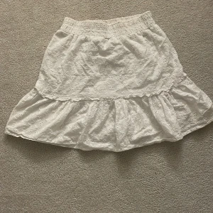 Vit volangkjol med broderade detaljer - Söt vit kjol med volang och broderade blommor. Resår i midjan för skön passform och luftig känsla. Perfekt för varma dagar och enkel att matcha med olika toppar.