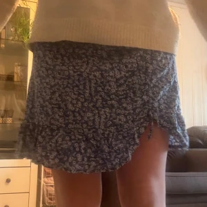 Blå blommig kortkjol med volang - Supersöt blå kortkjol med vitt blommönster och volangkant. Kjolen har en liten slits med knyt på sidan som ger en extra detalj. Perfekt för dig som gillar en feminin och somrig stil.
