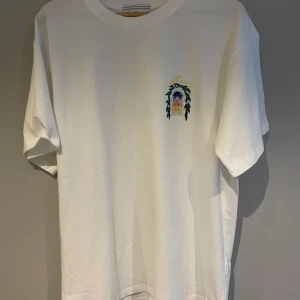 Vit t-shirt Casablanca - Säljer en vit t-shirt från Casablanca med ett färgglatt tryck av en byggnad och gröna blad på baksidan. Framsidan har ett mindre tryck med samma tema. Perfekt för en avslappnad stil.