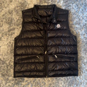 Svart dunväst från Moncler - Snygg svart dunväst från Moncler med quiltad design och två dragkedjefickor framtill. Västen har hög krage och klassisk Moncler-logga på bröstet. Perfekt för lager-på-lager under kyliga dagar. Nypris 9260kr.