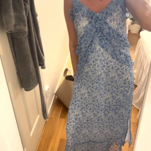 Blå blommig långklänning med volang - Säljer en  blå långklänning med blommigt mönster och volangdetaljer framtill. Klänningen är ärmlös och har en v-ringad halsringning.  säljer en på grund av att jag själv inte tyckte att jag passade i den! Så hoppas att någon annan kan nyttja den bättre än mig, den är ALDRIG ANVÄND💙✨ Köpt för : 500kr